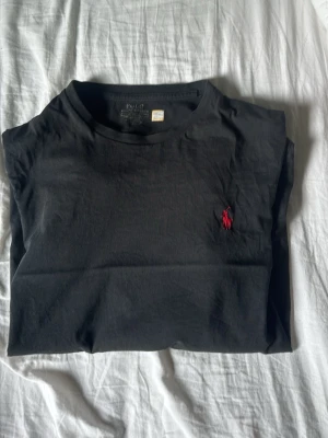 Svart t-shirt från Polo Ralph Lauren - Klassisk svart t-shirt från Polo Ralph Lauren med röd broderad logga på bröstet. T-shirten har rund hals och korta ärmar, perfekt till alla tillfällen. Tillverkad i mjuk bomull för skön känsla.