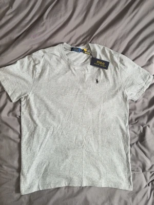 Grå t-shirt från Polo Ralph Lauren - Snygg grå t-shirt från Polo Ralph Lauren med klassisk passform och det ikoniska broderade logotypen på bröstet. Tillverkad i mjuk bomull som känns skön mot huden. Perfekt basplagg till garderoben och enkel att matcha med allt. STORLEK XXL i barn=L i herr
