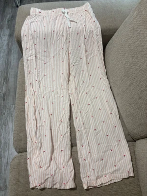 Randiga pyjamasbyxor med körsbär - Säljer ett par ljusrosa pyjamasbyxor från LCW Dream med vita ränder och små röda körsbärsmotiv. Byxorna har resår i midjan och en vit dragsko för extra komfort. Tillverkade i ett mjukt och lätt material, perfekta för mysiga kvällar hemma. Dessa är köpta utomlands och är i fel storlek, dessa har aldrig använts.