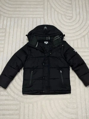 Svart pufferjacka från Burberry - Snygg svart pufferjacka från Burberry med huva och två dragkedjefickor framtill. Jackan har Burberry London England-tryck på ena ärmen och quiltad design. Perfekt för kalla vinterdagar och riktigt trendig look.