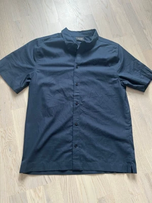 Mörkblå skjorta från Lyle & Scott - Snygg mörkblå skjorta från Lyle & Scott med korta ärmar och klassisk krage. Skjortan har knappar framtill och diskret logga på bröstet. Perfekt till sommarens alla tillfällen och enkel att matcha med jeans eller shorts.