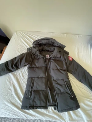 Svart dunjacka från Canada Goose - Säljer en svart dunjacka från Canada Goose med huva och dragkedja framtill. Jackan har två stora fickor med dragkedja och den klassiska Canada Goose-loggan på ärmen. Perfekt för kalla vinterdagar och riktigt varm tack vare dunfyllningen.
