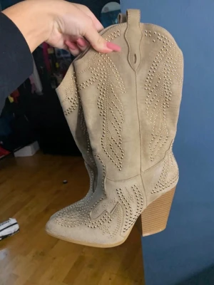 Beige cowboy boots med nitar - Snygga beige cowboy boots i mockaimitation med dekorativa nitar och klassiska broderidetaljer. Boots med hög klack och spetsig tå som ger en trendig westernkänsla. Perfekta för dig som vill sticka ut med din stil. Köpta för 1300