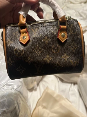  Monogram väska  - Superfin klassiskt brunt monogramcanvas med beige läderdetaljer och guldfärgade metalldelar. Väskan har två små handtag och dragkedja upptill. Perfekt för dig som gillar lyxiga accessoarer och ikonisk design.