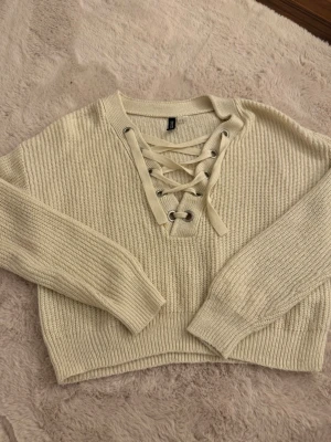 Beige stickad tröja med snörning - Säljer en beige stickad tröja med trendig snörning framtill och grov ribbstickning. Tröjan har långa ärmar, croppad passform och breda muddar vid ärmslut och nederkant. 