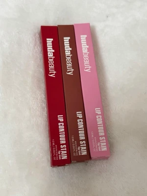 Huda Beauty Lip Contour Stain 3-pack - Säljer tre läppennor från Huda Beauty i färgerna Cherry Red, Warm Brown och Baby Pink. Pennorna har en klassisk smal form och kommer i snygga förpackningar som matchar färgerna. Perfekt för att skapa definierade och fylliga läppar.