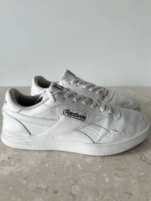 Vita Reebok sneakers i läder storlek 38  - Klassiska vita sneakers från Reebok i läder med lågt skaft och gröna loggor på hälen. Skorna har vita snören, diskreta sömmar och en stilren design som passar till det mesta. Perfekta för dig som gillar en clean och tidlös look.