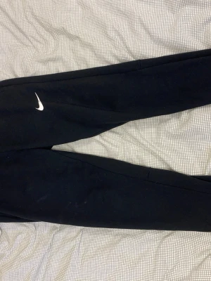 Svarta Nike sweatpants - Säljer ett par svarta sweatpants från Nike med klassisk vit Swoosh-logga på benet. Byxorna har resår i midjan och vid bensluten för en skön passform. Perfekta för träning eller chill. Tillverkade i mjukt material för extra komfort.
