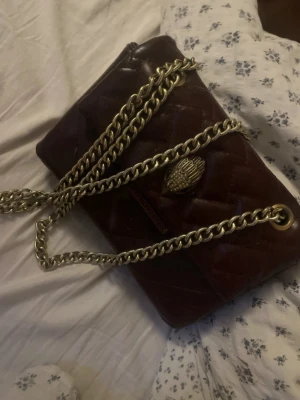 Vinröd axelväska från Kurt Geiger - Snygg vinröd axelväska från Kurt Geiger i quiltat läder med guldfärgad kedjerem och ikonisk örndetalj på locket. Väskan har en elegant rektangulär form och guldfärgad logoplatta på baksidan. Perfekt accessoar för att lyfta din outfit.