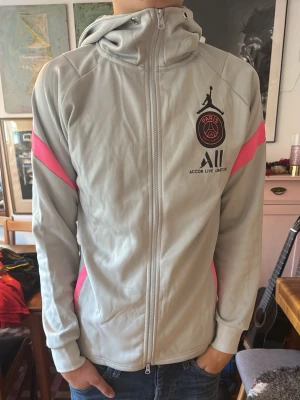 PSG x Jordan grå hoodie med dragkedja - Snygg grå hoodie från Jordan x PSG med dragkedja framtill och rosa detaljer på ärmarna. Stor PSG-logga och Jumpman-logga på bröstet samt texten 'Accor Live Limitless'. Tillverkad i mjukt syntetmaterial och har huva. Köpt begagnat och äkthet kan ej garanteras