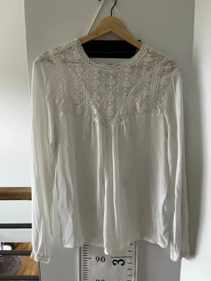 Vit blus med spetsdetaljer - Säljer en vit blus med långa ärmar och vackra spetsdetaljer över axlar och bröst. Blusen har en lös passform och tunna, luftiga material som ger en romantisk vibe. Perfekt till jeans eller kjol för en trendig look. Andrig använd, köpt second hand