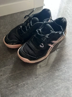 Svarta Asics sneakers med rosa detaljer - Säljer ett par svarta Asics sneakers med rosa detaljer och vit logga på sidan. Skorna har en låg profil, snörning och ovandel i mesh och syntetmaterial. Sulan är ljusrosa och ger en sportig look. Perfekta för dig som gillar stilrena och bekväma sneakers.