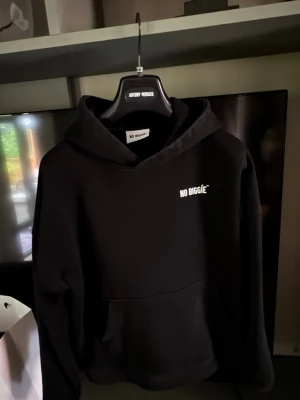 Svart hoodie från No Biggie - Säljer en svart hoodie från No Biggie med vit logga tryckt på bröstet och stort tryck på ryggen. Hoodien har en klassisk känguruficka framtill och en rymlig huva. Perfekt för dig som gillar streetwear och enkel stil.