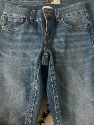 Blå jeans från Gina Tricot - Säljer ett par klassiska blå jeans från Gina Tricot i storlek 170. Jeansen har fem fickor, normalhög midja och snygga slitningar för en trendig look. Tillverkade i mjukt denimtyg med kontrastsömmar och bälteshällor.