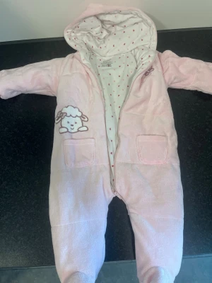 Rosa overall med får från Walking - Supersöt rosa overall med huva från Walking, dekorerad med ett broderat får på bröstet och små fickor framtill. Insidan är vit med små hjärtan i rött och orange. Tillverkad i 80% bomull och 20% polyester. Perfekt för mysiga dagar!