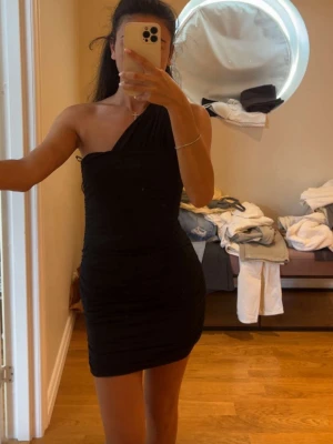 Svart oneshoulder kort klänning - Snygg svart kort klänning med oneshoulder-design. Klänningen har en tight passform och är perfekt för dig som vill ha en stilren och trendig look. Materialet känns mjukt och följsamt mot kroppen.