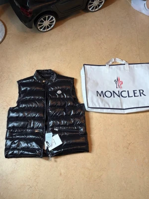 Svart dunväst från Moncler - Snygg svart dunväst från Moncler med glansig finish och klassisk logotyp på bröstet. Västen har hög krage, dragkedja framtill och två stora fickor med dragkedja. Perfekt för lager-på-lager och streetstyle.