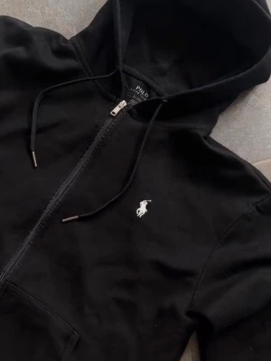 Svart hoodie från Polo Ralph Lauren - Snygg svart hoodie med dragkedja från Polo Ralph Lauren. Klassisk design med vit broderad logga på bröstet, huva med dragsko och kängurufickor framtill. Tillverkad i mjukt bomullsmaterial som är skönt att ha på sig.