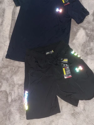 Svart träningsset från Under Armour - Snyggt svart träningsset från Under Armour med t-shirt och shorts. Båda plaggen har regnbågsfärgade loggor och är tillverkade i ett lätt och snabbtorkande material, perfekt för gymmet eller löprundan. Shortsen har dragsko i midjan och tryckt text på benet.