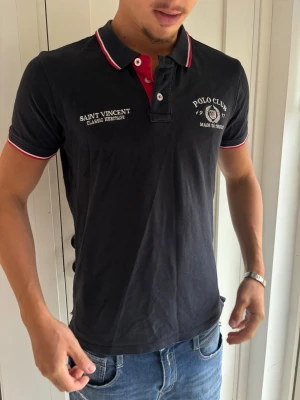 Svart pikétröja med tryck - Svart pikétröja med vita och röda detaljer på krage och ärmslut. Tryck på bröstet med texten 'Polo Club' och 'Saint Vincent Classic Heritage'. Kortärmad modell med knappslå framtill.
