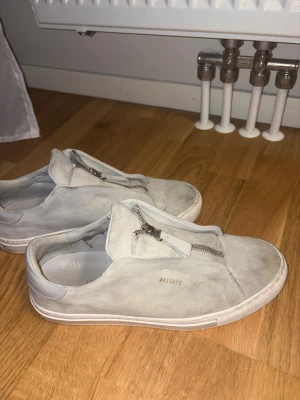 Ljusa sneakers från Axel Arigato - Snygga ljusgrå sneakers från Axel Arigato i mocka med dragkedja framtill istället för klassiska snören. Skorna har en låg profil och vit sula, samt diskret logga på sidan. Perfekta för dig som gillar minimalistisk och stilren design.