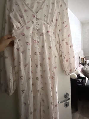 Lång blommig klänning med knappar - Romantisk långklänning i ljus färg med rosa blommigt mönster. Klänningen har v-ringning, knappar framtill och långa ärmar. Perfekt för sommarens alla tillfällen. Materialet känns lätt och luftigt.