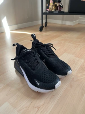 Nike Air Max 270 svarta sneakers - Säljer ett par svarta Nike Air Max 270 sneakers med vit sula och synlig Air-enhet i hälen. Skorna har mesh-överdel för bra ventilation, klassisk Nike-logga på sidan och snörning. Perfekta för dig som gillar sportig och modern stil.