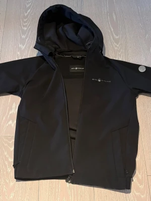 Svart softshell vindjacka Sail Racing - Svart softshell vindjacka från Sail Racing med huva och dragkedja framtill. Jackan har två sidofickor med dragkedja och diskret logga på bröstet. Materialet är 94% polyester och 6% spandex, vilket ger en stretchig och bekväm känsla.