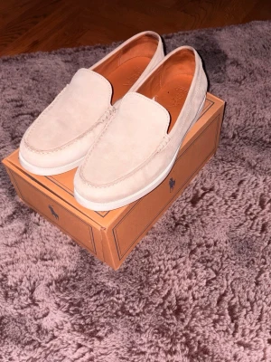 Beige loafers från Polo Ralph Lauren - Stilrena beige loafers i mocka från Polo Ralph Lauren. Skorna har en klassisk slip-on design med vit sula och diskreta sömmar runt ovandelen. Perfekta för en avslappnad men snygg look.använd ett fåtal gånger 2-4. Ny pris 2295