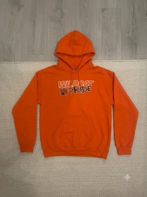 Orange hoodie Wildcat Pride Gildan - Orange hoodie från Gildan med trycket 'Wildcat Pride' och tassavtryck på bröstet. Klassisk modell med huva, dragsnören och stor magficka. Tillverkad i mjuk bomullsblandning, perfekt för chill dagar eller skolan.