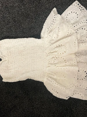 Vit broderad sommarklänning - Supersöt vit klänning med smockad överdel och volangig kjol i broderad bomull. Klänningen har ett vackert hålmönster och är ärmlös, perfekt för varma dagar. Materialet känns lätt och luftigt, och den har en romantisk och trendig look. Den är väldig stretchig och har inbyggda shorts, använd en gång 