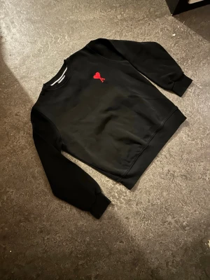 Svart sweatshirt från Ami Paris - Svart sweatshirt från Ami Paris med röd broderad logga på bröstet. Klassisk rund halsringning och ribbade muddar vid ärmslut och nederkant. Tillverkad i mjukt bomullsmaterial för en skön och avslappnad look.