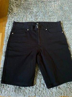 Svarta jeansshorts från H&M - Säljer ett par svarta jeansshorts från H&M med klassisk femficksmodell och knappgylf. Shortsen har en rak passform och bälteshällor i midjan. Perfekta för sommaren och enkla att matcha med olika stilar.