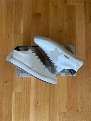 Golden Goose Super-Star Sneakers - Snygga vita Super-Star sneakers från Golden Goose med svart häl och grå stjärndetalj på sidan. Skorna har grå skosnören och en chunky sula. Tillverkade i läder med klassisk låg profil och diskreta håldetaljer på sidan.