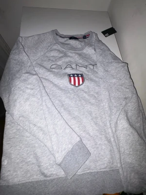 Grå sweatshirt från GANT - Snygg grå sweatshirt från GANT med broderad logga och ett sköldmärke på bröstet. Tröjan har rund halsringning, ribbade muddar vid ärmslut och nederkant samt en klassisk relaxed fit. Tillverkad i mjukt bomullsmaterial som är skönt mot huden.