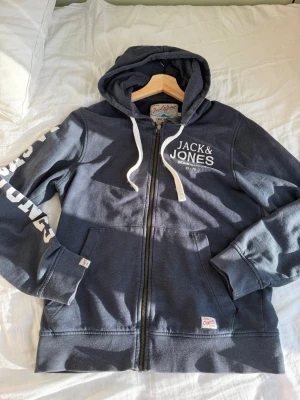 Jack & Jones hoodie - Mörkblå hoodie med dragkedja från Jack & Jones. Stor vit logga på bröstet och tryck på ena ärmen. Utrustad med huva och vita dragsnören samt två fickor framtill. Tillverkad i mjukt bomullsmaterial för en skön och avslappnad stil.