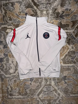 Vit PSG träningsjacka med röd detalj - Snygg vit träningsjacka från PSG x Jordan med hel dragkedja framtill. Jackan har röda ränder på axlarna, PSG-logga på bröstet och Jordan-logga i svart. Perfekt för dig som gillar sportig stil och fotbollsinspirerade plagg.