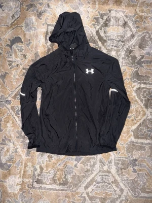 Svart vindjacka från Under Armour - Snygg svart vindjacka från Under Armour med huva och hel dragkedja framtill. Jackan har reflekterande detaljer på bröstet och ärmen samt elastiska muddar. Tillverkad i lätt och tunt material som passar perfekt för blåsiga dagar.