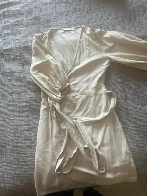 Beige omlottklänning med knytband - Snygg beige omlottklänning från NA-KD med långa puffärmar och knytband i midjan. Klänningen har v-ringning och är tillverkad i ett mjukt linneliknande material. Perfekt för en trendig och avslappnad look.