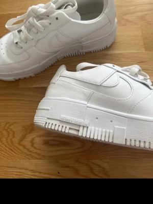 Vita Nike Air Force 1 sneakers med kubformig  - Fräscha helvita Nike Air Force 1 sneakers med klassisk siluett och chunky sula. Skorna har perforerad tå, vita skosnören och diskret Nike-logga på sidan. Tillverkade i läder med detaljer i gummi för extra hållbarhet och stil.