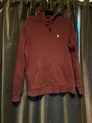 Vinröd hoodie med gul logga - Säljer en vinröd hoodie med huva och känguruficka framtill. På bröstet sitter en liten gul broderad logga. Hoodien har ribbade muddar vid ärmslut och nederkant samt dragsko i huvan. Perfekt för dig som gillar enkel men snygg stil.