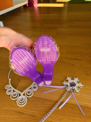 Prinsessetillbehör lila set - Säljer ett prinsessetillbehör-set med lila och silverfärgade detaljer. Innehåller ett par lila plastklackskor med dekorativa rosetter, ett gnistrande halsband, en tiara och en trollstav med pärlor och lila hjärta. Perfekt för maskerad eller cosplay.