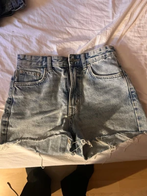 Högmidjade ljusblå jeansshorts - Snygga högmidjade jeansshorts i ljusblå denim med råa, fransiga kanter. Klassisk femficksmodell med dragkedja fram. Perfekta för varma dagar och enkel att matcha med olika toppar.