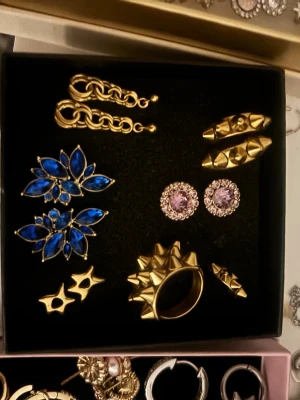 Mix av örhängen och ringar i guld och blått - Säljer ett set med flera örhängen och ringar i guldton. Inkluderar chunky kedjeörhängen, blå stenar i blommig form, lila glittriga studs, samt ringar med nitar och stjärndetaljer. Perfekt för dig som vill mixa och matcha accessoarer. Man kan köpa sakerna separat så kom med ett pris förslag på vad du vill ha 