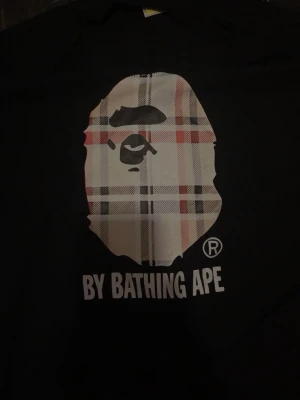 Svart BAPE t-shirt med rutig logga - Säljer en svart t-shirt från BAPE med den ikoniska apansiluetten i rutigt mönster på bröstet och texten 'BY BATHING APE' under. Skön passform och perfekt för dig som gillar streetwear och vill sticka ut.