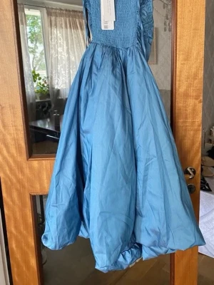 Blå långklänning med puffig kjol - Säljer en blå klänning med puffig kjol och smockad överdel. Klänningen har ärmlös design och en elegant, voluminös siluett som ger en riktigt snygg look. Perfekt för dig som vill sticka ut med en unik och trendig stil.