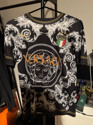 Italy fotbollströja - Säljer en svart t-shirt med vita barockinspirerade mönster och guldiga detaljer från Italien . Stort Medusa-tryck framtill, guldfärgad text och italiensk flaggpatch på bröstet. Klassisk rund halsringning med grekiskt mönster. Perfekt statement-plagg!