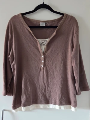 Beige topp med spetsdetalj och knappar - Säljer en beige topp med trekvartsärm och v-ringning. Toppen har dekorativa knappar framtill och en vit spetsdetalj vid halsen samt vit kant nertill. Perfekt för dig som gillar en avslappnad men söt stil. Hittar inget märke.
