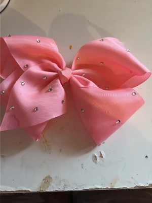 Rosa hårrosett med strass - Stor rosa hårrosett i ribbat tyg dekorerad med glittrande strasstenar. Perfekt accessoar för att ge frisyren extra bling och färg. Rosetten har en klassisk form och är lätt att fästa i håret. Har en fläck se bild! 