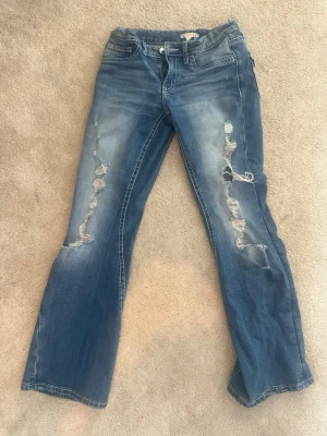 Blå slitna bootcut jeans från Gina 14+ - Säljer ett par blå bootcut jeans med slitningar och hål på framsidan av båda benen. Jeansen har klassisk femficksdesign och ljusare tvättade partier. Perfekta för en avslappnad och trendig look. Low waist och justerbara band i midjan så de passar dig perfekt!